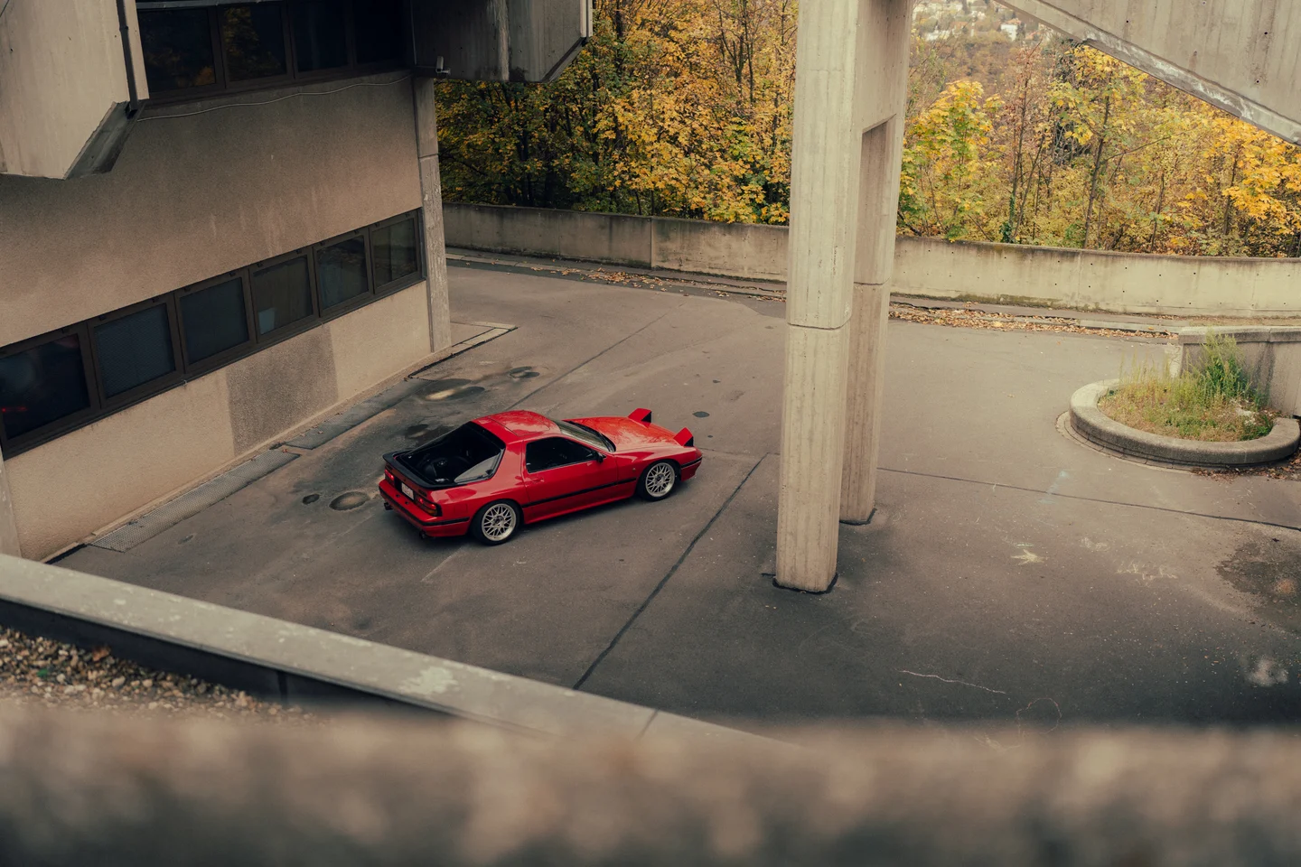 A red Mazda RX-7 FC3S beneath concrete walkways, surrounded by autumn trees.